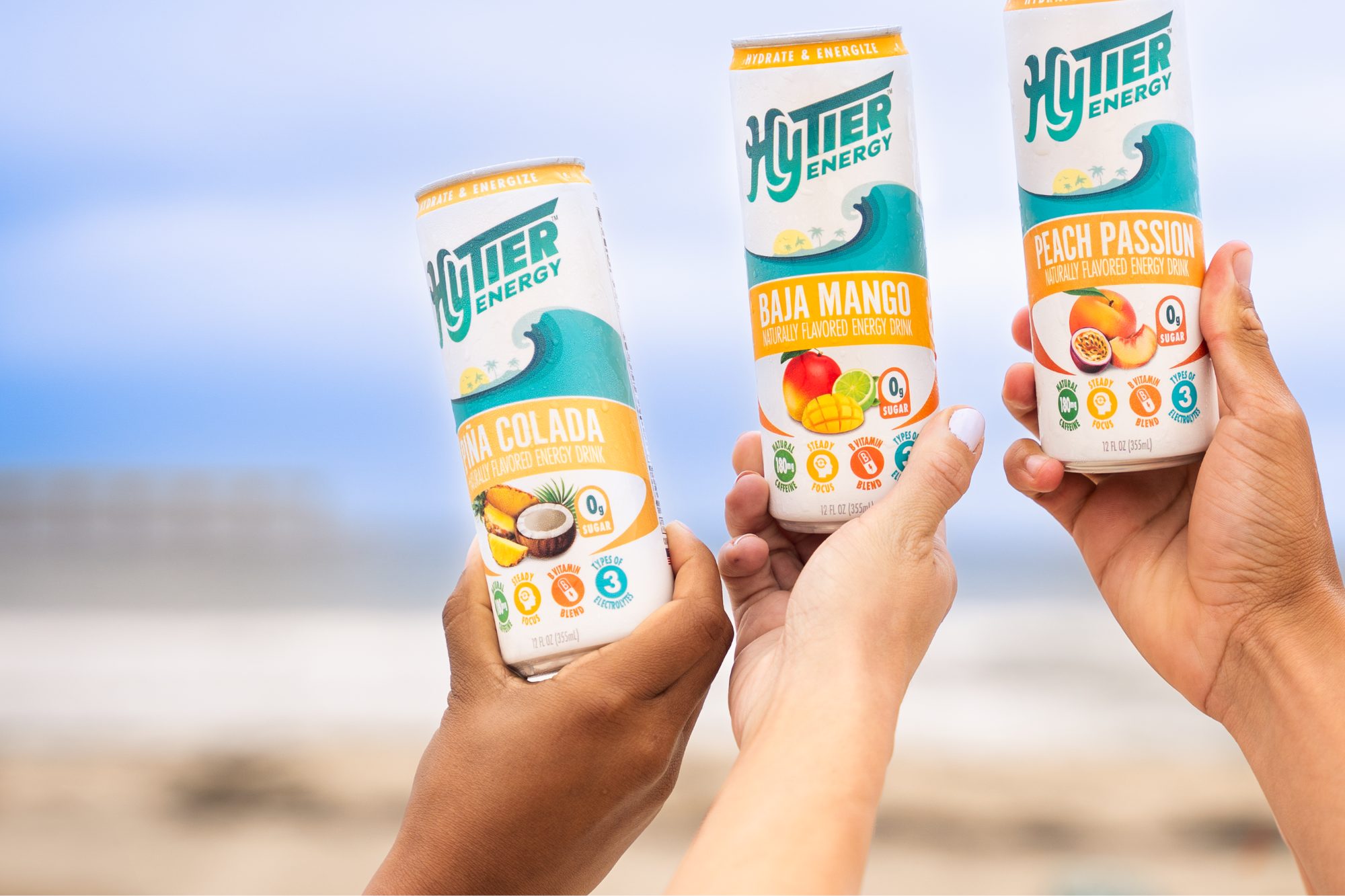Hands raising HyTier Energy cans in Piña Colada, Baja Mango and Peach Passion against a blue sky
